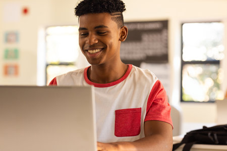 Teenage boy smiling while working on laptop at classroom desk, with black backpack and chalkboard....