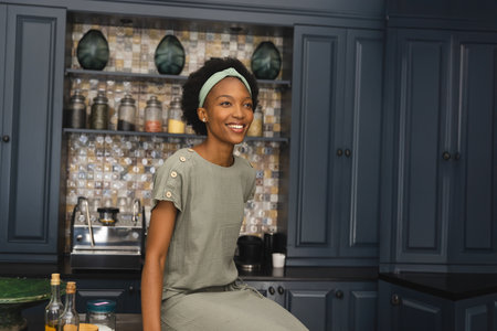 African American woman sitting on kitchen island, surrounded by oil bottles and mosaic backsplash. Modern, bright, vibrant, eclectic, open shelving, culinary, residentialの写真素材