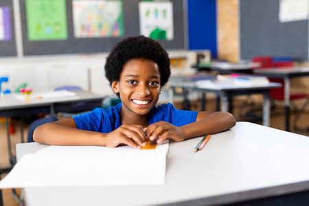 African American boy sitting at elementary classroom desk writing in open notebook with pencils....