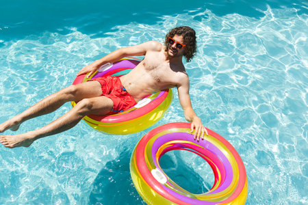 Young man floating on multicolored inflatable ring in outdoor swimming pool, holding second ring....