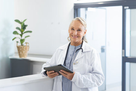 Senior female doctor using tablet at clinic reception desk beside potted plant, wearing stethoscope....