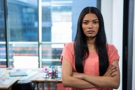 Woman standing before cluttered studio desk with papers, crossing arms near windows with blinds. Professional, creative, modern, office, productivity, workflow, brainstormingの写真素材