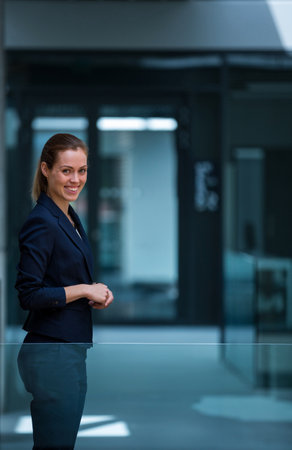 Standing woman wearing navy blue suit leaning near glass railing in office corridor, copy space. Professional, corporate, modern, sleek, confident, businesswear, workplaceの写真素材