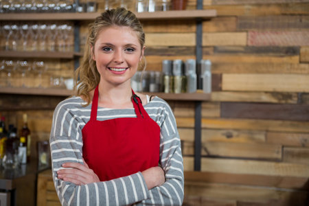 Smiling woman wearing red apron standing in cafÃ©, with wooden shelves, glassware and spice jars. Hospitality, barista, rustic, cheerful, beverage, culinary, kitchenの写真素材
