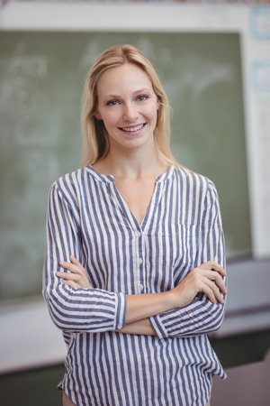 Smiling female teacher standing in classroom, with green chalkboard showing faint writing. Education, training, mentorship, instruction, learning, professional, academicの写真素材