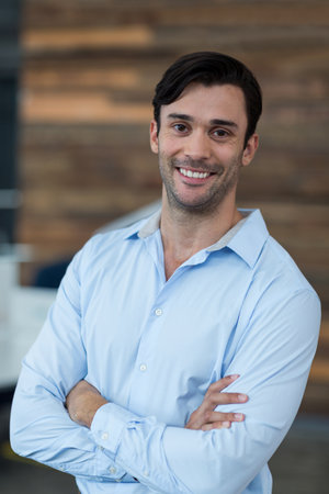 Man standing with crossed arms and smiling in modern office interior, with wooden accent wall. Professional, teamwork, corporate, modern, productivity, success, businessの写真素材