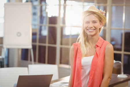 Smiling business woman wearing hat standing by male colleague looking at laptop in creative officeの写真素材