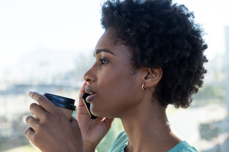 African American woman talking on smartphone holding coffee cup in office near window, copy space....