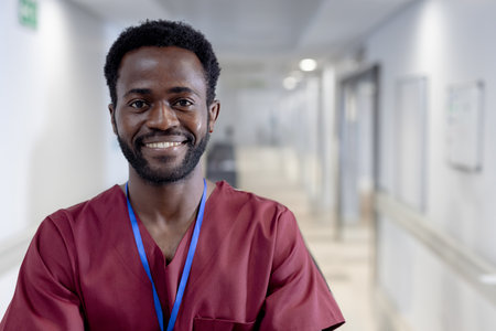 African American male standing in hospital hallway wearing maroon scrubs, blue lanyard badge. Nurse,...