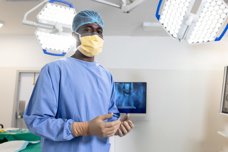 African American male surgeon donning gloves and mask in operating room with surgical lights....