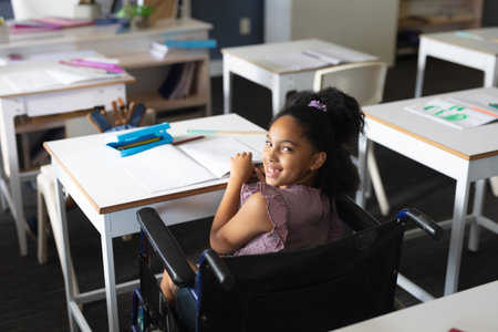 Young girl rolling in wheelchair in classroom, smiling with blue folder and pencil on desk. Education, accessible, classroom, positivity, engagement, learning, diversityの写真素材