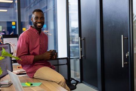 African American man sitting on desk in office, holding coffee mug with open laptop notebook....
