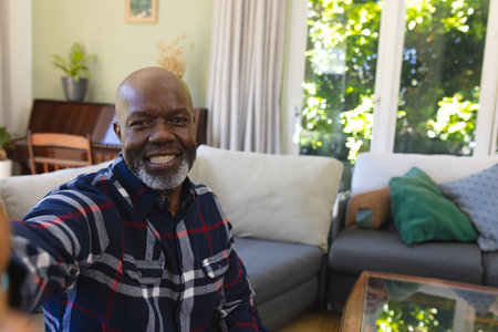 Senior African American man taking selfie with smartphone on sofa in living room, copy space. Cozy,...