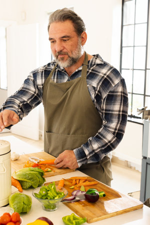 Senior man chopping carrots on wooden cutting board at home kitchen island, with fresh vegetables. Cooking, meal prep, culinary, nutrition, wellness, ingredients, produceの写真素材