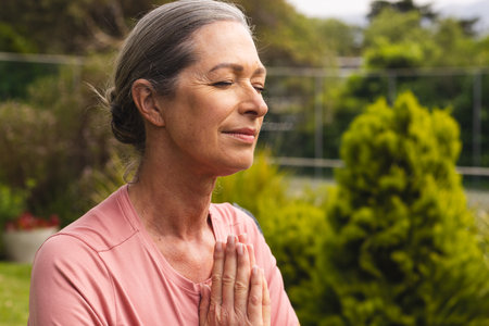 Senior woman meditating with hands clasped in backyard garden under sunlight, copy space. Mindfulness, contemplation, spirituality, wellness, serenity, calm, outdoorsの写真素材