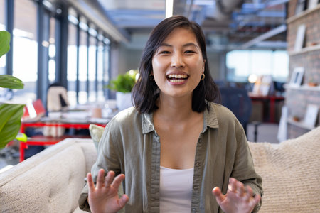 Asian woman smiling and gesturing toward camera in office lounge, with beige sofa, potted plant....