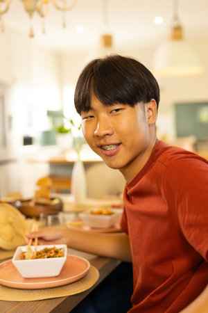 Young Asian man eating noodles from square bowl with chopsticks at table in modern dining room....