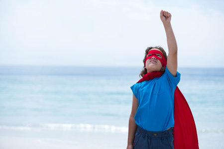 Child striking heroic pose on sandy shoreline with calm ocean, wearing red superhero mask and cape. Adventure, determination, confidence, fantasy, outdoor, vibrant, youthfulの写真素材