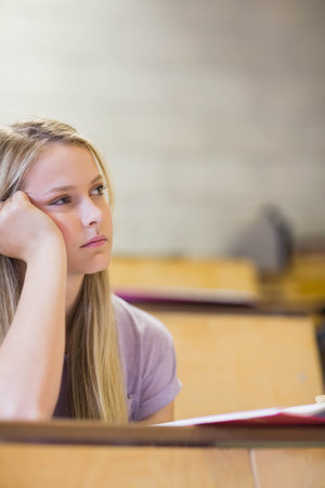 Teenage girl resting head on hand gazing at wooden classroom desk with red notebook, copy space. Student, education, study, classroom, concentration, learning, academicの写真素材