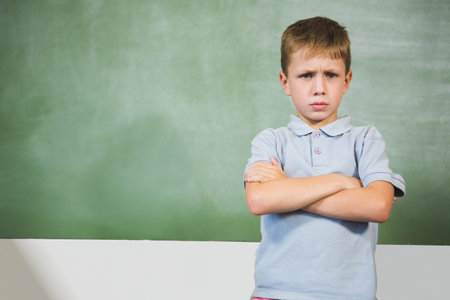 Boy student standing with folded arms and frowning at green chalkboard in classroom, copy space. Contemplative, educational, introspective, mood, serious, focused, classroomの写真素材