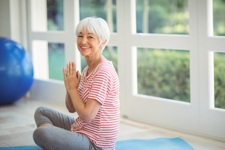 Senior woman wearing workout clothes sitting on blue mat beside exercise ball at home near windows. Wellness, stability, serenity, natural light, tranquility, mindfulness, agingの写真素材