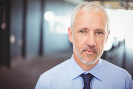 Mature man standing wearing light blue shirt and navy tie in office corridor with blurred columns. Professional, corporate, leadership, modern, executive, formal, business environmentの写真素材