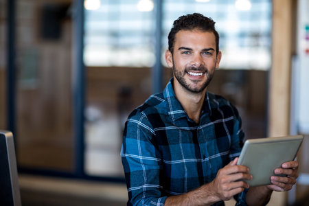 Bearded man wearing plaid shirt sitting at desk in office meeting room holding tablet while smiling....