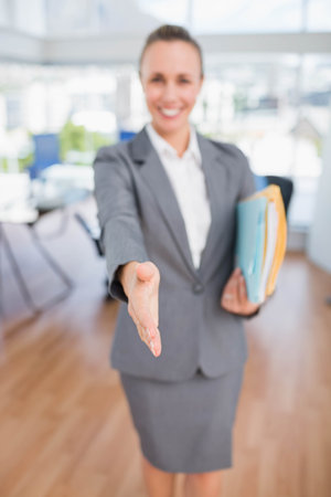 Female business professional in blazer extending right hand in greeting in office with file folders. Corporate, networking, professional, workspace, modern, leadership, communicationの写真素材