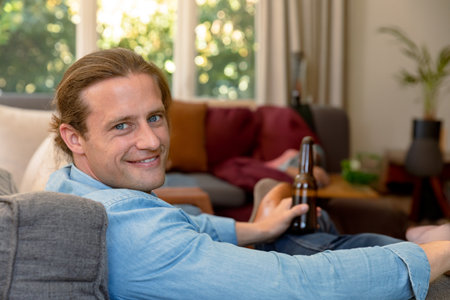 Male reclining on gray sofa in living room with potted plant, holding dark beer bottle. Relaxation, comfort, cozy, domestic, casual, serene, modernの写真素材