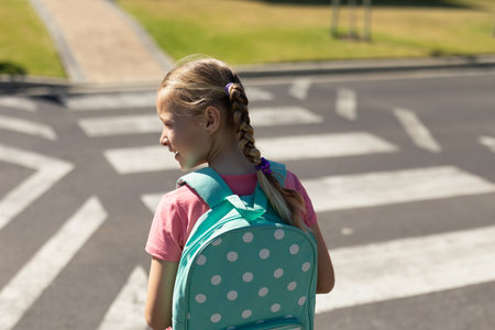 Schoolgirl standing on zebra crossing at suburban street, carrying mint-green polka-dot backpack. Children, urban, casual, neighborhood, youthful, outdoors, safetyの写真素材