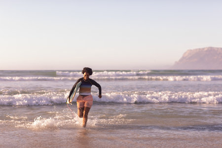 African American woman running from surf toward sandy shore holding surfboard wearing rash guard....