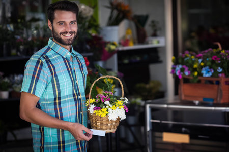 Man in late twenties standing inside flower shop holding wicker flower basket and blank card. Florist, floral, vibrant, retail, botanical, natural, arrangementの写真素材