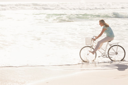 Woman riding white bicycle with front basket skimming surf along sandy beach barefoot, copy space. Scenic, leisure, adventure, coastal, outdoor, serene, vibrantの写真素材