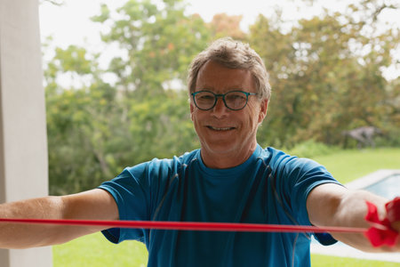 Senior man wearing teal shirt stretching red resistance band on patio overlooking lawn and pool....