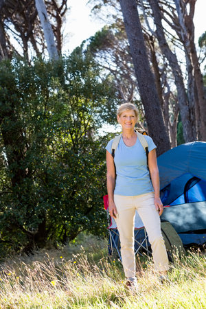 Senior woman standing in forest campsite wearing hiking backpack beside blue tent and camping chair. Outdoors, adventure, nature, relaxation, discovery, exploration, scenicの写真素材