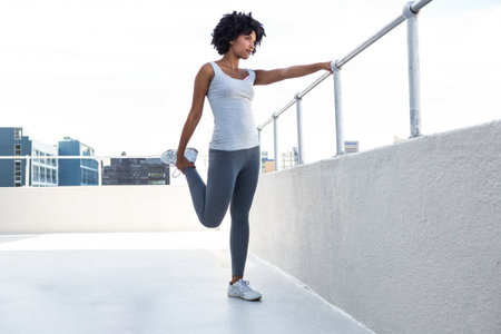 African American woman performing quad stretch on rooftop terrace using metal railing and sneakers....