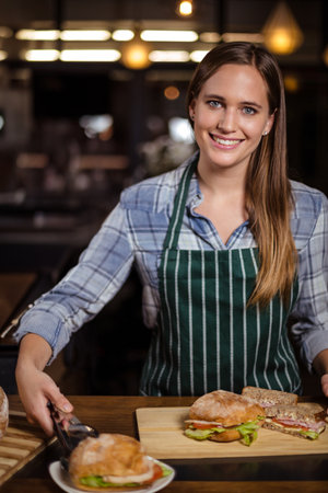 Female barista standing behind wooden counter assembling sandwiches in cafÃ© under pendant lights....