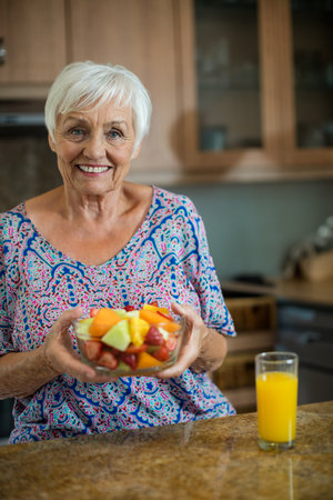 Senior woman sitting at home kitchen counter holding glass bowl of fruit salad and orange juice. Healthy lifestyle, wellness, freshness, nutrition, cozy, vibrant, naturalの写真素材