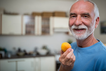 Senior man standing in kitchen holding yellow apricot near mouth, wearing blue t-shirt, copy space. Health, wellness, nutrition, lifestyle, natural, rustic, homelyの写真素材