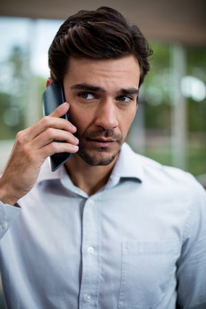 Middle-aged man standing near windows in modern office holding smartphone to ear wearing blue shirt....