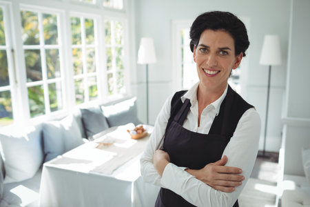 Woman standing smiling in bright breakfast nook wearing black apron over white shirt, copy space. CafÃ©, hospitality, warmly lit, inviting, professional, interior design, comfortの写真素材