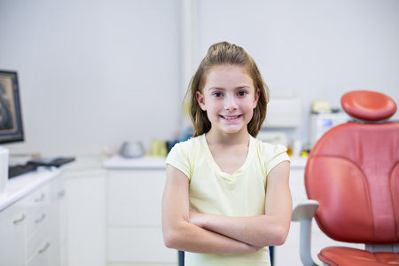 Female child standing with arms crossed in dental clinic with red dental chair and computer monitor. Pediatric, healthcare, clinical, hygiene, bright, cheerful, medicalの写真素材