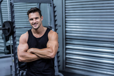 Muscular man wearing black tank top standing at gym by squat rack with plates, copy space. Strength, fitness, training, athletic, motivation, industrial, dynamicの写真素材