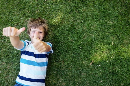 Child lying on sunlit park lawn wearing striped tee and shorts giving thumbs up, copy space. Playfulness, leisure, outdoor, happiness, brightness, innocence, relaxationの写真素材