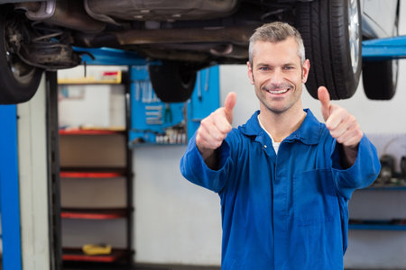 Male mechanic standing under lifted car on lift in garage with toolbox giving thumbs-up, copy space. Automotive, repair, industry, workshop, maintenance, tools, vehicleの写真素材