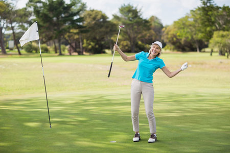 Female golfer in golf attire holding putter and celebrating on green with flagstick, copy space. Athletic, victory, sport, leisure, outdoor, competitive, energeticの写真素材
