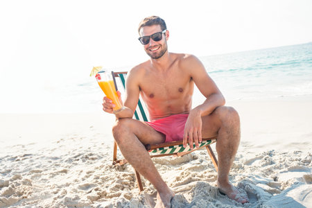 Man in red swim shorts, sunglasses sitting on beach chair at shore holding cocktail umbrella straw. Vacation, leisure, relaxation, seaside, lifestyle, tropical, outdoorの写真素材