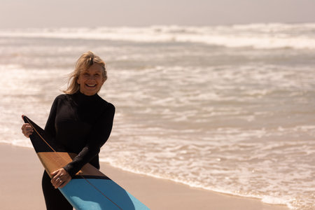 Senior woman holding wooden-nose turquoise-base surfboard in black wetsuit on shore, copy space. Adventure, resilience, outdoor, freedom, active, leisure, harmonyの写真素材