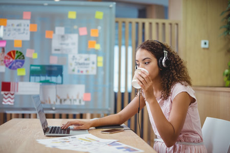 Woman wearing light pink dress and headphones, sipping coffee, reviewing charts on laptop at office....