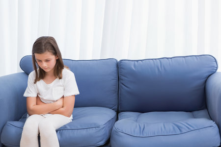 Female child sitting on blue sofa with cushions in living room near white curtains, copy space. Youth, comfort, serenity, contemplation, interior, casual, pastelの写真素材